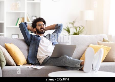 Relaxed Young Indian Man With Laptop Sitting On Couch At Home Interior Stock Photo