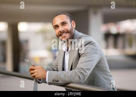 Pensive Young Businessman With Smartphone In Hands Standing Outdoors And Looking Away Stock Photo