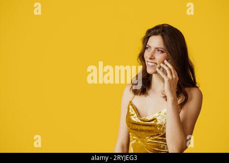 Young caucasian lady in elegant dress talking on cellphone and smiling, looking at free space on yellow background Stock Photo