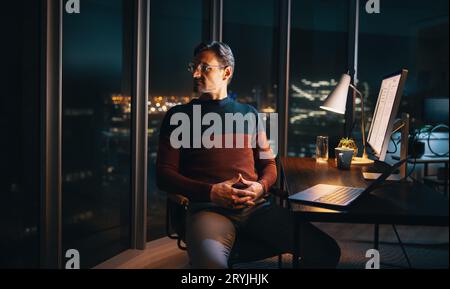 A pensive caucasian businessman working late in the office. He sits at his desk, engrossed in his laptop, looking away into the night, thinking about Stock Photo