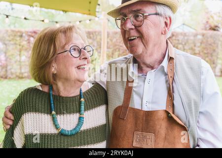 Elderly Couple Embracing at Outdoor Gathering - A loving elderly couple in their 60s to 70s embrace outdoors with a festively lit tent in the backgrou Stock Photo