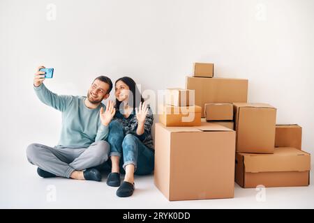 Happy smiling couple greeting friends at online video call using smartphone app sitting on floor surrounded by cardboard boxes Stock Photo