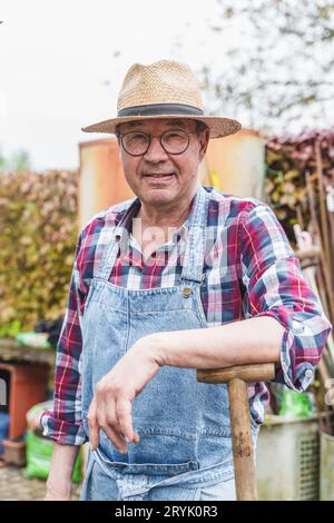 Elderly Farmer Leaning on His Hoe - Portrait of a 65-year-old farmer, wearing glasses and a straw hat, leaning on his hoe, showcasing years of experie Stock Photo
