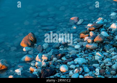Colorful pebbles on the blue river shore, photographed at the last minutes of golden sunset, colouring some of the stones in orange Stock Photo