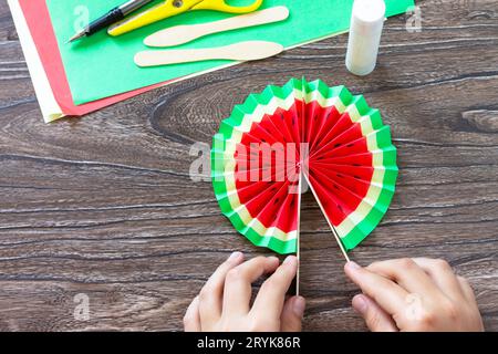 In the hands of a child Paper Fan watermelon on a wooden table. Childrens art project, handmade, crafts for children. Stock Photo