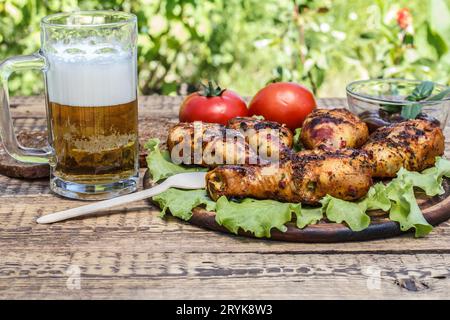 Grilled chicken legs and lettuce leaves on wooden chopping board, tomatoes and glass mug of beer. Stock Photo