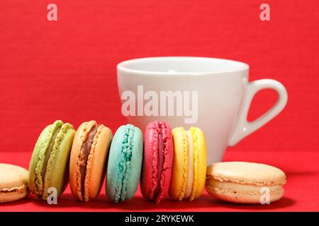 Sweet macarons cakes of different colors with a cup on the red textured surface. Stock Photo