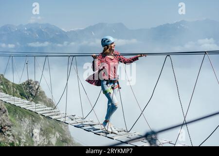 A happy cheerful woman walks on a suspension bridge high in the mountains. Wanderlust and adventures. Extreme vacation. Stock Photo