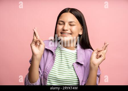 Dreaming smiling teenage girl making wish crossing fingers with closed eyes on pink background. Stock Photo