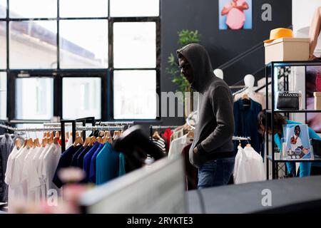 African american man wearing hood and glasses trying to steal fashionable clothes from modern boutique. Robber looking around to see if someone watching him while stealing merchandise Stock Photo