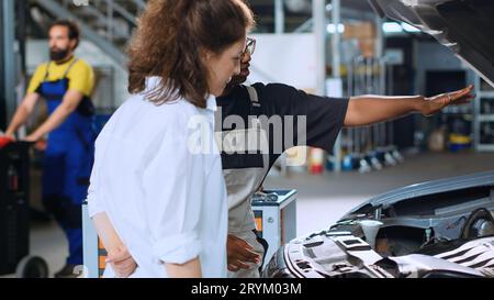 Helpful engineer assisting customer with car maintenance in auto repair shop. Licensed expert in garage looking over car components with woman, servicing her vehicle during annual checkup Stock Photo