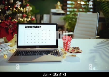 modern Christmas decorated green office with december calendar on laptop screen, poinsettia, red cup of hot Christmas beverage and cookies. Stock Photo
