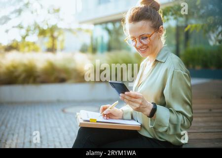 smiling modern female employee near office building in green blouse and eyeglasses with smartphone, documents and folder. Stock Photo