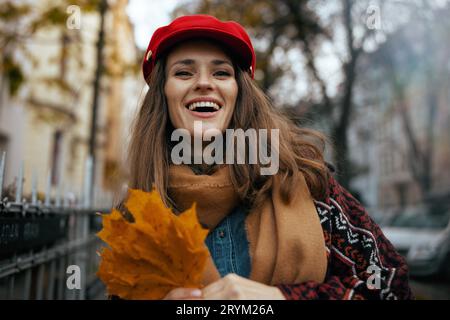 Hello autumn. Portrait of happy trendy 40 years old woman in red hat with autumn leafs and scarf in the city. Stock Photo