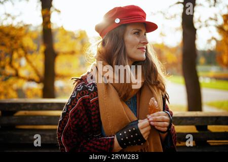 Hello autumn. pensive trendy middle aged woman in red hat with autumn leaf, scarf and gloves sitting on bench in the city park. Stock Photo