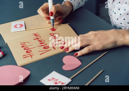 Woman writes in a sketch book with a felt-tip pen Happy Valentine's Day. Attributes for Valentine's Day on a gray desktop. Stock Photo