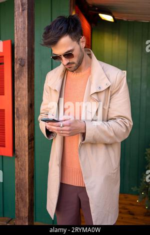 Fashionable man in coat chatting outside. Elegant dark haired male reading text message on his phone. Online communication: handsome man texting with cell phone during Christmas holidays Stock Photo