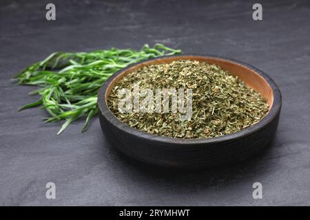 Dry tarragon in bowl and green leaves on black textured table, closeup Stock Photo
