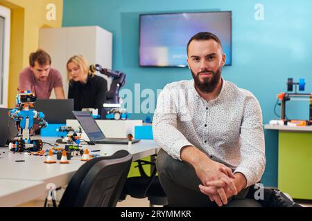 A man sitting in a robotics laboratory while his colleagues in the background test new, cutting edge robotic inventions. Stock Photo