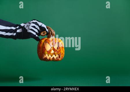 Skeleton gloved hand holds ceramic pumpkin jack-o'-lantern on green background Stock Photo
