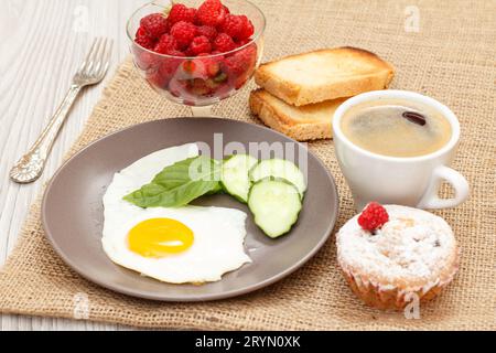 Plate with fried egg, cut cucumber and leaf of basil, muffin, cup of black coffee, toasts Stock Photo