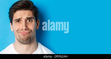 Head shot of skeptical and disappointed man, grimacing complicated, standing against blue background Stock Photo