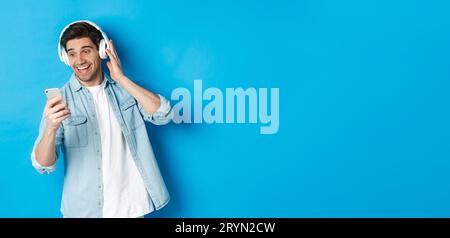 Happy man listening to music in headphones and reading message on smartphone, smiling excited, standing against blue background Stock Photo