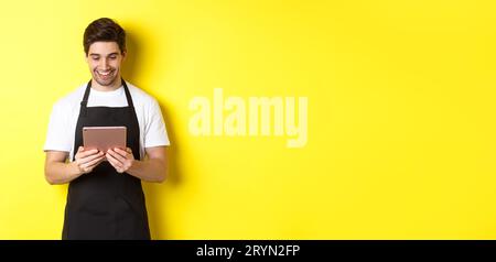 Seller in black apron looking at digital tablet screen, smiling pleased, standing over yellow background Stock Photo