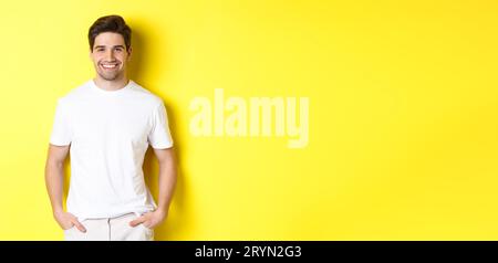 Young handsome man smiling at camera, holding hands in pockets, standing against yellow background Stock Photo