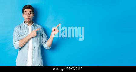 Portrait of surprised bearded guy in casual outfit pointing fingers right, showing amazing promo offer, standing over blue backg Stock Photo