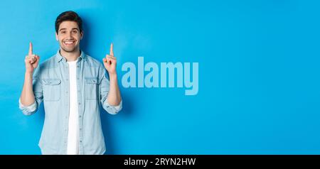 Portrait of handsome bearded man in casual outfit, smiling happy and pointing fingers up at copy space, standing over blue backg Stock Photo