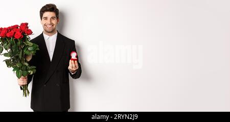 Handsome smiling man in black suit, holding roses and engagement ring, making a proposal to marry him, standing against white ba Stock Photo