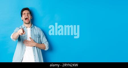 Adult man in casual outfit laughing out loud and pointing at you, looking at something funny, chuckling while standing against b Stock Photo