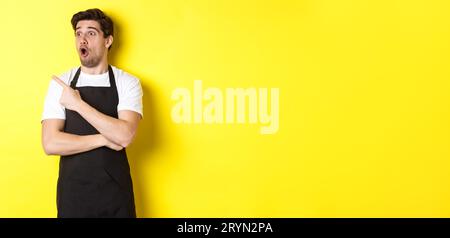 Waiter in black apron checking out promo offer, pointing finger and looking left at your logo, standing over yellow background Stock Photo