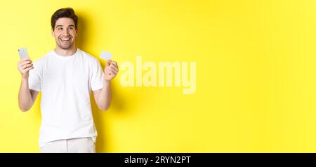 Happy man thinking about shopping, holding credit card and smartphone, smiling pleased, standing over yellow background Stock Photo
