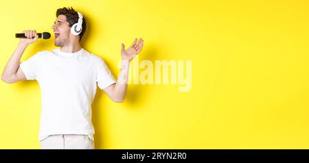 Passionate guy in headphones holding microphone, singing karaoke song, standing over yellow background in white clothes Stock Photo