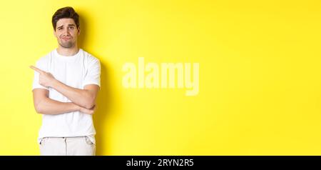 Skeptical and reluctant guy in white t-shirt, pointing finger left and smirking unamused, showing bad advertisement, standing ov Stock Photo