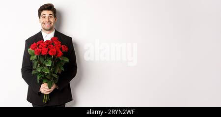 Image of handsome man in black suit, holding bouquet of roses and smiling, standing against white background Stock Photo