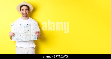 Travelling, vacation and tourism concept. Smiling young man going on trip, holding road map and smiling, standing over yellow ba Stock Photo