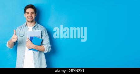 Young man student with notebooks, showing thumb up in approval, smiling satisfied, blue studio background Stock Photo
