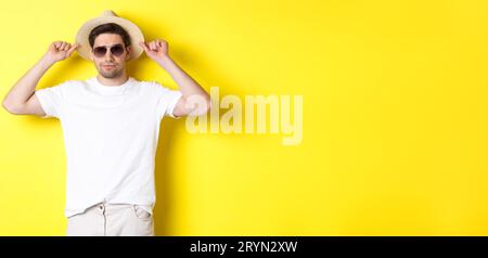 Confident young male tourist ready for vacation, wearing straw hat and sunglasses, standing against yellow background Stock Photo
