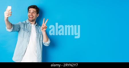 Happy man taking selfie and showing peace sign on blue background, holding mobile phone Stock Photo