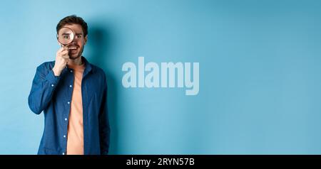 Cheerful young man found something good, smiling and looking through magnifying glass, standing on blue background Stock Photo