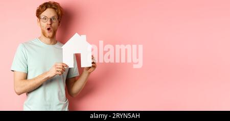 Real estate. Surprised young man with red hair and beard, wearing glasses and t-shirt, showing paper house cutout and looking im Stock Photo