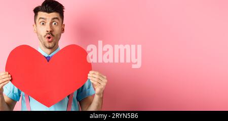 Valentines day concept. Amused young man with bow-tie and moustache, showing heart cutout and looking for love, standing on pink Stock Photo