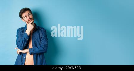 Pensive young man touching face and looking aside at empty space, imaging something, thinking off plan, standing on blue backgro Stock Photo