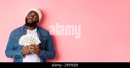 Dreamy african-american man thinking about shopping, holding money dollars and looking upper left corner, standing over pink bac Stock Photo