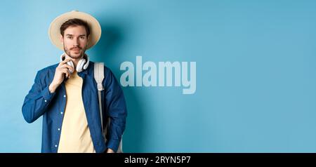 Handsome guy travelling with headphone, wearing straw hat and smiling, standing with backpack on blue background Stock Photo