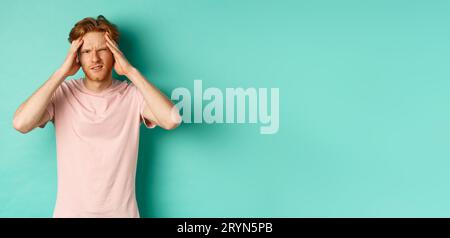 Young redhead man touching head and looking dizzy, feeling headache or migraine, standing in t-shirt over mint background Stock Photo