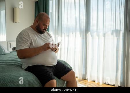 Fat bearded bald guy with smartphone chatting, dials number, types message, watches news, weather Stock Photo